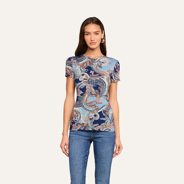 A woman wearing the Ressi Fitted Tee—a slim fit, short sleeve top in a midnight blue paisley pattern—paired with blue jeans, stands against a plain light background facing forward with her arms relaxed by her sides.