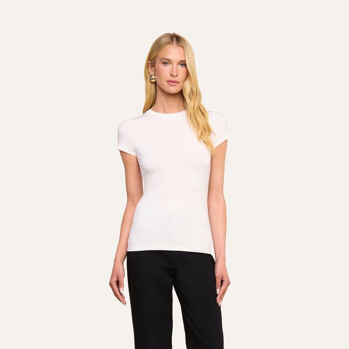 A woman with long blonde hair wears the Ressi Fitted Tee—a form-fitting, plain white crew neck top—paired with black pants, standing against a light neutral background.