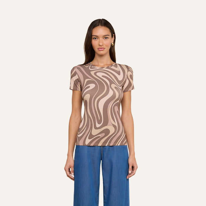 A woman stands against a light background wearing the Ressi Fitted Tee—a short-sleeve, beige and brown abstract swirl print top in stretch jersey knit—paired with blue jeans. She has long, straight brown hair and faces forward with a neutral expression.