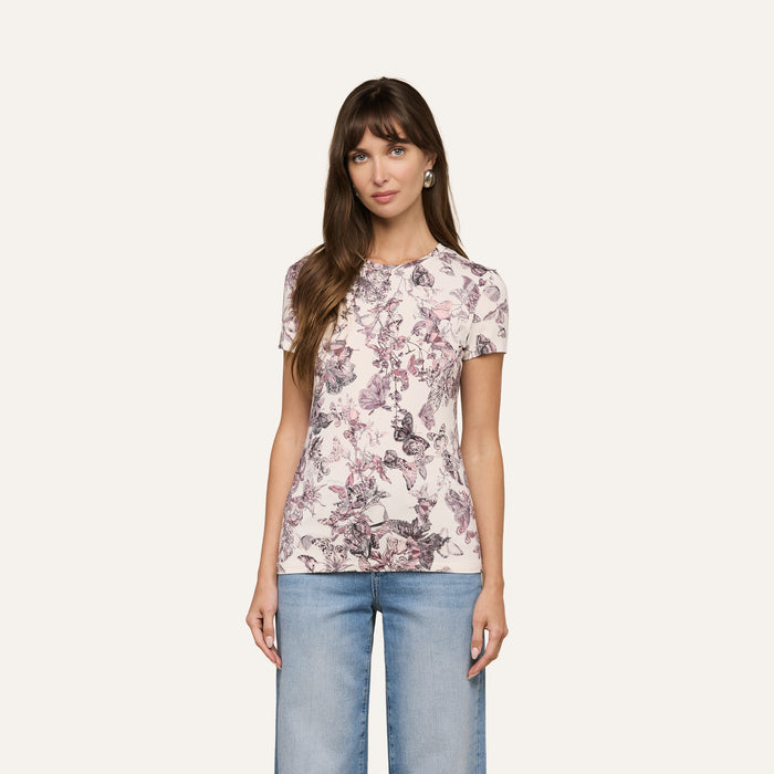 A woman wears the Ressi Fitted Tee, a pink floral slim cut short-sleeve shirt, paired with light blue jeans. She stands against an off-white background, looking at the camera with her arms relaxed at her sides.