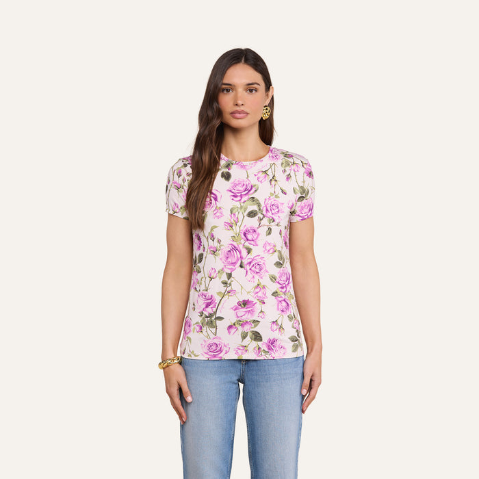 A woman wears the bestselling Ressi Fitted Tee in white with a pink and purple floral print, styled with light blue jeans, gold hoop earrings, and a gold bracelet, standing against a plain off-white background.