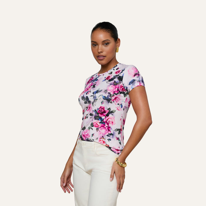 A woman poses wearing the Ressi Fitted Tee, a bestselling top crafted from stretch jersey knit. The tee showcases a pink rose print with purple and white floral accents. She pairs it with white pants and gold jewelry, set against a plain light background.