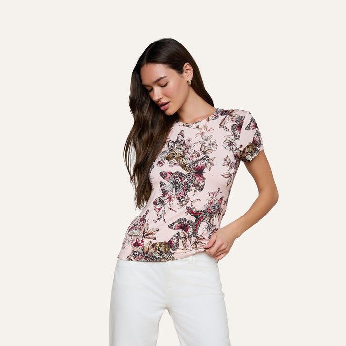 A woman with long brown hair is wearing the Ressi Fitted Tee—a light pink, floral-patterned stretch jersey knit shirt—paired with white pants. She stands against a plain background, looking down with one hand in her pocket.