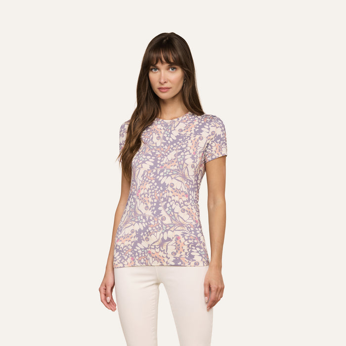 Woman with long brown hair wearing the bestselling Ressi Fitted Tee, featuring short sleeves and a pastel floral pattern, paired with white pants and standing against a plain light background.