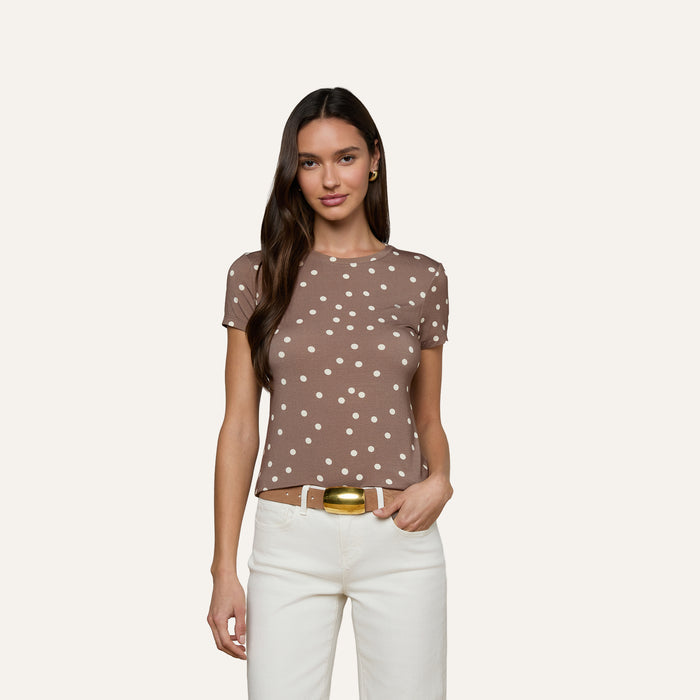 A woman with long brown hair wears the Ressi Fitted Tee, a slim-cut brown short-sleeve shirt with white polka dots, paired with white pants. She stands against a plain light background, one hand in her pocket.