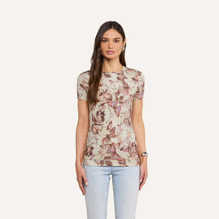 A woman with long brown hair and a neutral expression stands against a plain background, wearing the bestselling Ressi Fitted Tee—crafted from stretch jersey and featuring an intricate butterfly and floral print—paired with light blue jeans.