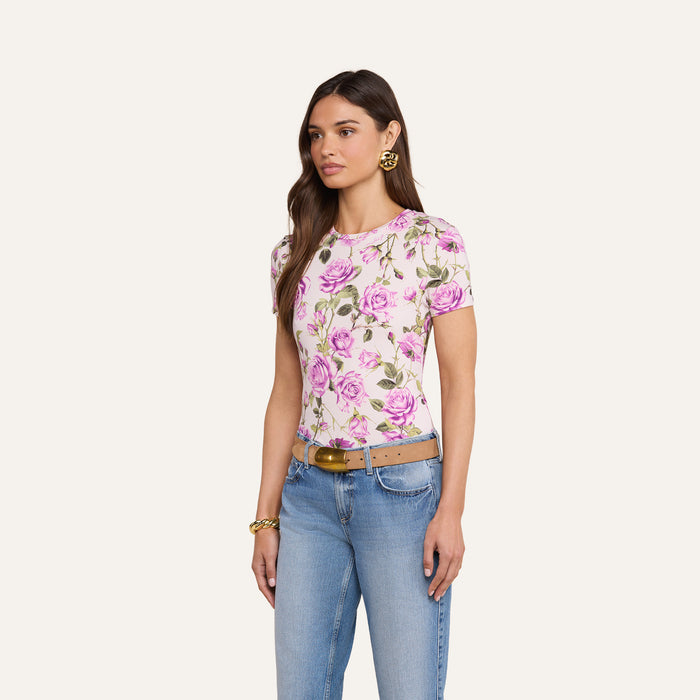 A woman is wearing the bestselling Ressi Fitted Tee in white with a pink floral print, paired with light blue jeans, a tan belt, gold hoop earrings, and a gold bracelet, standing against a plain light background.