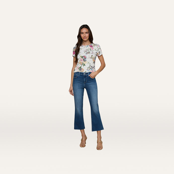 A woman stands against a plain background wearing the Ressi Fitted Tee in white butterfly garden print, blue cropped flare jeans, and beige heeled sandals.