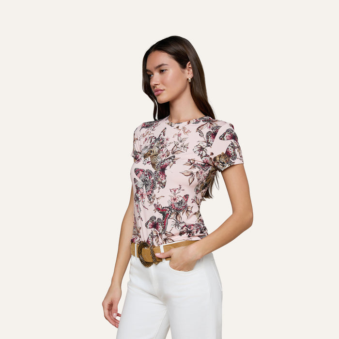 Woman wearing the bestselling Ressi Fitted Tee in light pink with a floral pattern, made from stretch jersey knit, paired with white pants, standing with one hand in her pocket against a plain light background.