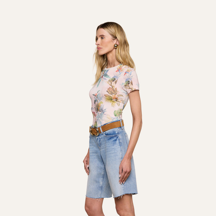 Woman standing in profile against a light background, wearing the Ressi Fitted Tee—a slim-cut pastel floral short-sleeve top—tucked into knee-length denim shorts with a tan belt and gold hoop earrings.
