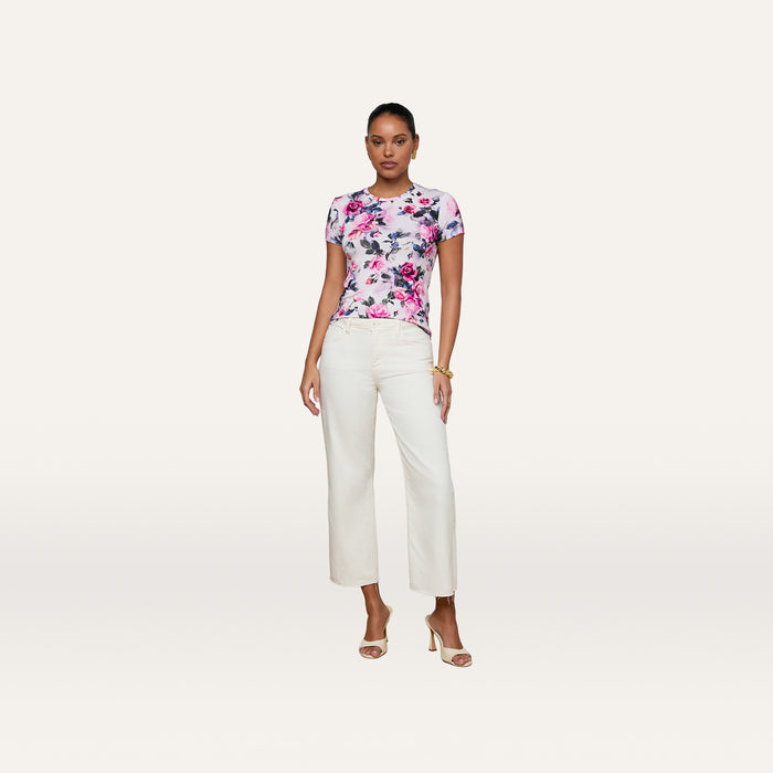 A woman stands against a white background, wearing the Ressi Fitted Tee in a short-sleeve pink rose print stretch jersey knit, paired with white wide-leg pants, nude high-heeled sandals, and gold bracelets.