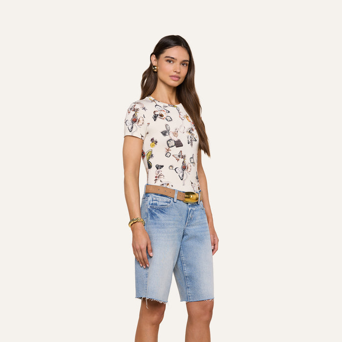 A woman with long brown hair wears the bestselling Ressi Fitted Tee—a white, slim-cut t-shirt featuring a butterfly print—paired with light blue knee-length denim shorts, a brown belt, and gold jewelry, standing against a plain light background.