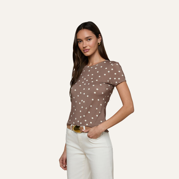 A woman poses against a plain light background in the bestselling Ressi Fitted Tee, a brown short-sleeve top with white polka dots crafted from stretch jersey knit and designed for a slim fit. She pairs it with white pants, her long hair down, standing with one hand on her hip and a neutral expression that highlights the figure-flattering silhouette of the tee.