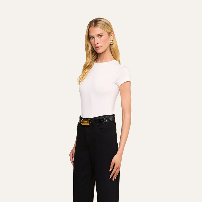 A woman with long blonde hair stands against a plain light background, wearing the Ressi Fitted Tee in white, paired with black high-waisted pants, a black belt with a gold buckle, and gold earrings.