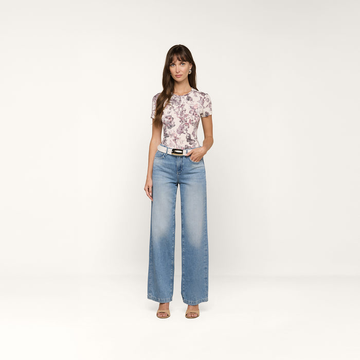 A woman stands against a plain white background wearing the bestselling Ressi Fitted Tee styled as a floral short-sleeve top, paired with light blue wide-leg jeans, a black belt, and open-toed shoes. She has long brown hair with bangs and her left hand is in her pocket.