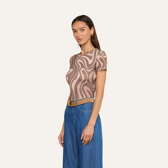 Woman wearing the Ressi Fitted Tee, a short-sleeve stretch jersey knit top with an abstract swirl print in brown and beige, paired with blue wide-leg jeans and a brown belt, standing against a plain off-white background.