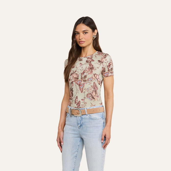 A woman stands against a plain background, wearing the bestselling Ressi Fitted Tee with a pink floral pattern, paired with light blue jeans and a beige belt. The Ressi Fitted Tee features short sleeves, a slim cut, and is crafted from soft stretch jersey. She has long brown hair and a neutral expression.