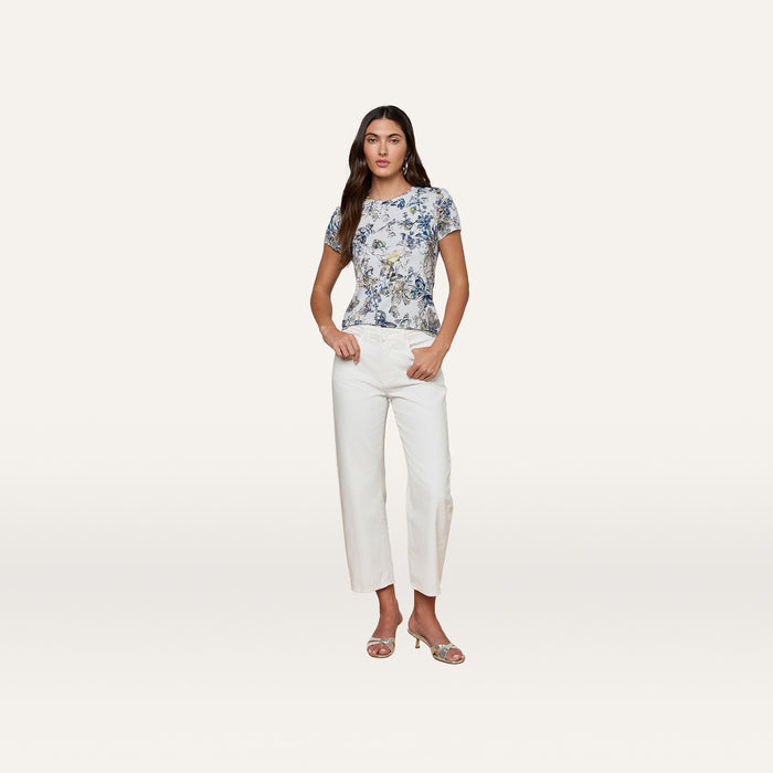 A woman stands against a plain white background, wearing the Ressi Fitted Tee—a floral short-sleeve stretch jersey knit top—paired with white pants and open-toed heels, her hands in her pockets.