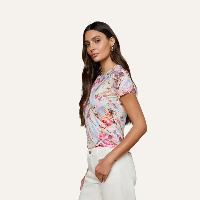 A woman with long brown hair stands sideways against a plain light background, wearing the bestselling Ressi Fitted Tee in a pastel print stretch jersey knit and white pants.