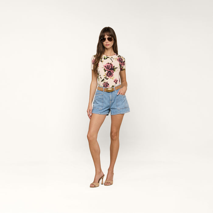 A woman wearing sunglasses, the bestselling Ressi Fitted Tee with a slim floral short-sleeve style, denim shorts with a belt, and high heels stands against a plain white background.