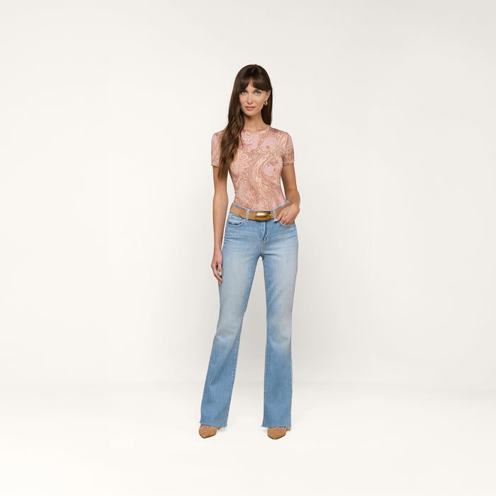 A woman stands against a plain white background, wearing the Ressi Fitted Tee in a mauve paisley print, paired with light blue jeans, a brown belt, and tan heels. She faces forward with a relaxed expression and her arms by her sides.