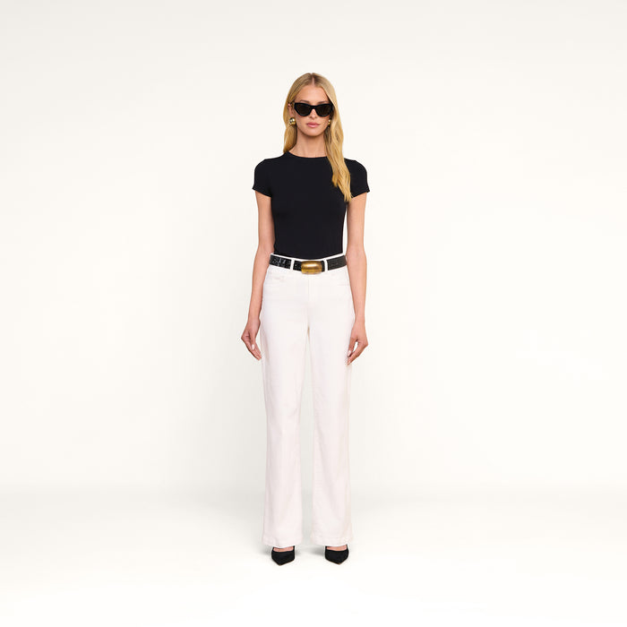 A woman stands against a plain white background wearing the Ressi Fitted Tee, styled with white high-waisted pants, black heels, a black belt with a gold buckle, sunglasses, and gold earrings.