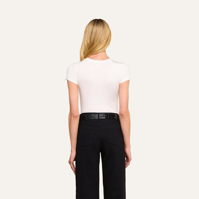 A person with long blonde hair is standing with their back to the camera, wearing the form-fitting Ressi Fitted Tee in white, paired with black pants and a black belt against a plain light background.