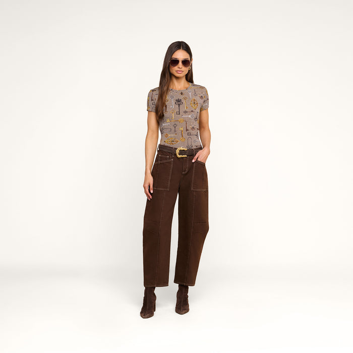 A woman wearing sunglasses, the Ressi Fitted Tee with short sleeves, slim cut brown pants with patch pockets, and brown boots stands against a plain white background with one hand in her pocket.