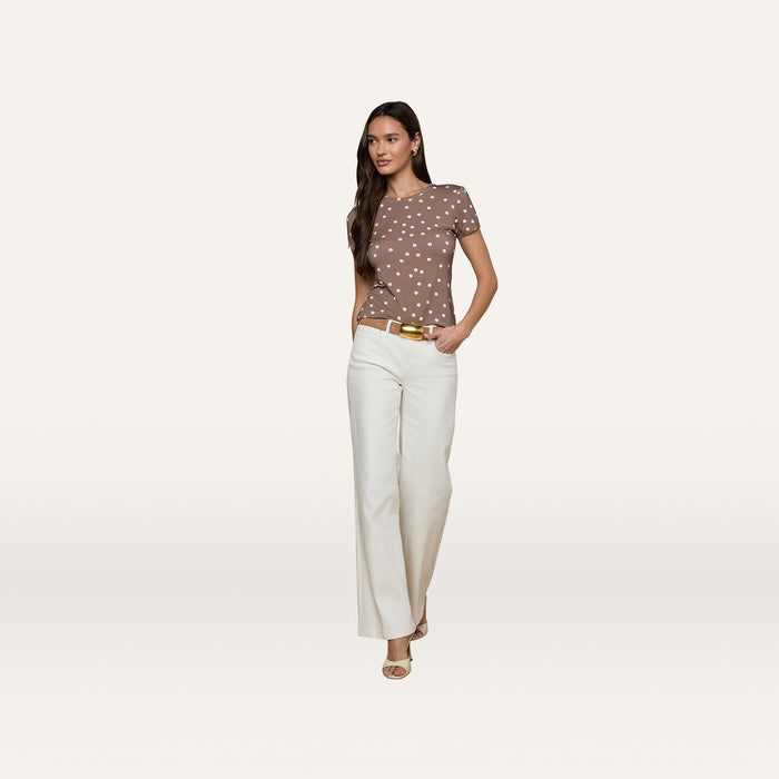 A woman stands against a plain white background, wearing the Ressi Fitted Tee—a brown short-sleeve polka dot top crafted from stretch jersey knit—paired with white wide-leg pants and beige heels, with one hand in her pocket.