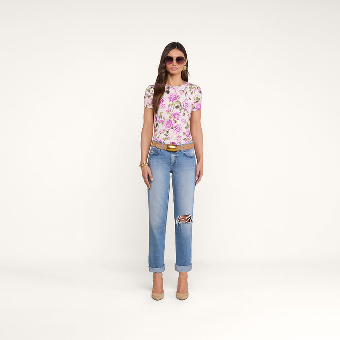 A woman with long brown hair stands against a plain white background wearing sunglasses, the pink floral Ressi Fitted Tee, light blue ripped jeans, beige heels, and gold accessories. Her hands are at her sides.