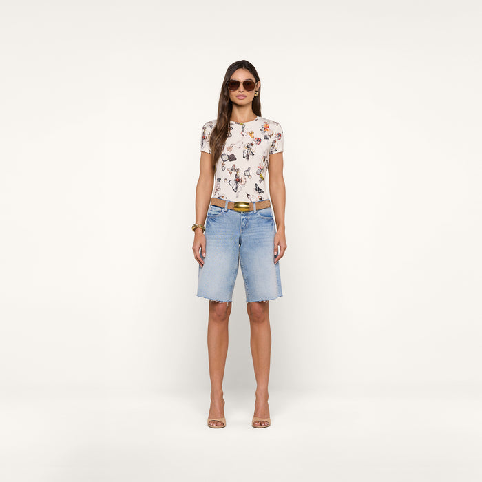 A woman stands facing forward, wearing sunglasses, the bestselling Ressi Fitted Tee featuring a floral graphic, light blue slim-cut knee-length denim shorts, a brown belt, and beige heels against a plain white background.