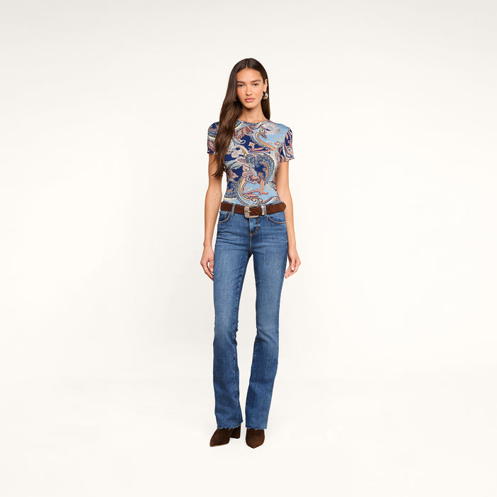 A woman stands facing forward against a plain white background, wearing the Ressi Fitted Tee in a midnight blue paisley print, paired with blue jeans, a brown belt, and brown shoes.