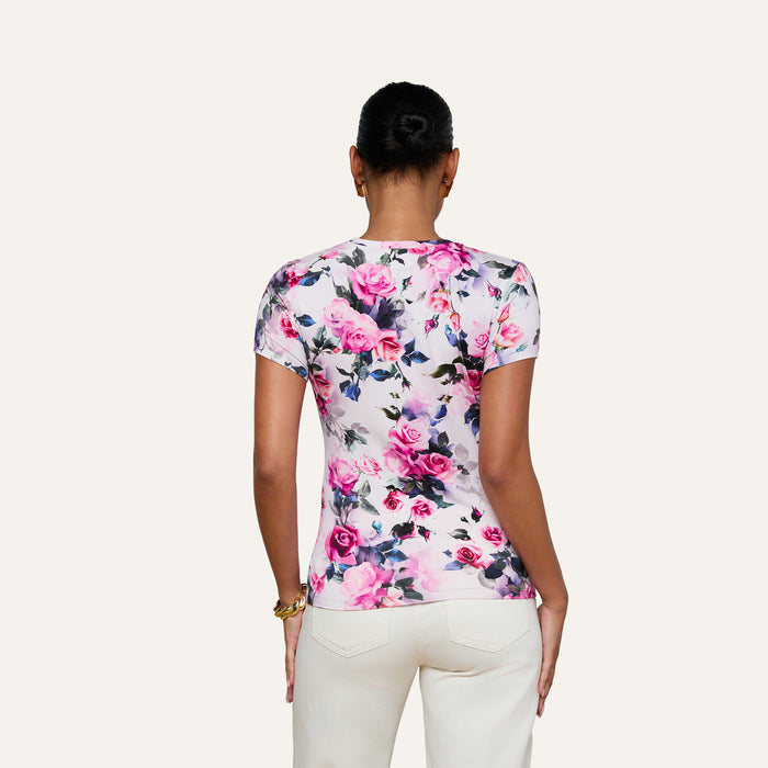 A woman stands with her back to the camera, wearing the bestselling Ressi Fitted Tee in a stretch jersey knit with a pink rose print, paired with white pants. Her hair is styled in a neat bun against a plain light background.