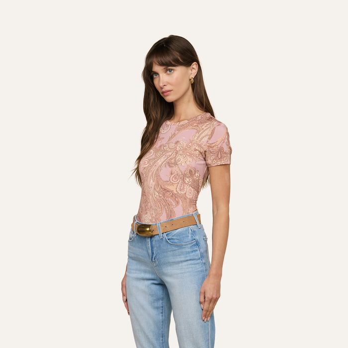 A woman with long brown hair is wearing the Ressi Fitted Tee—a short-sleeve mauve paisley print stretch jersey knit top—paired with blue jeans and a tan belt, standing against a plain light background.