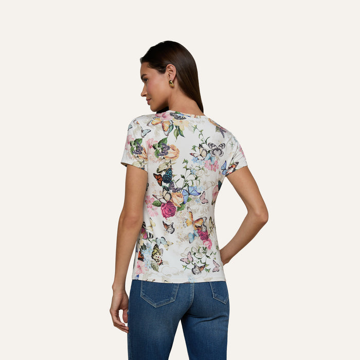 A woman stands with her back to the camera, wearing the Ressi Fitted Tee—bestselling and adorned with a floral and butterfly garden print—paired with blue jeans against a plain light background.