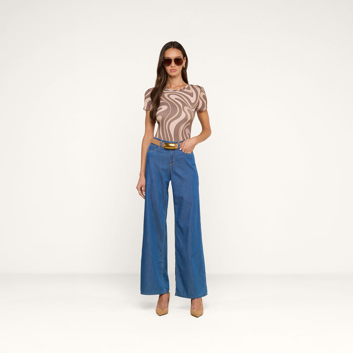 A woman wearing sunglasses, the Ressi Fitted Tee—an abstract swirl print beige and brown patterned top—wide-leg blue jeans with a belt, and tan heels stands against a plain white background.