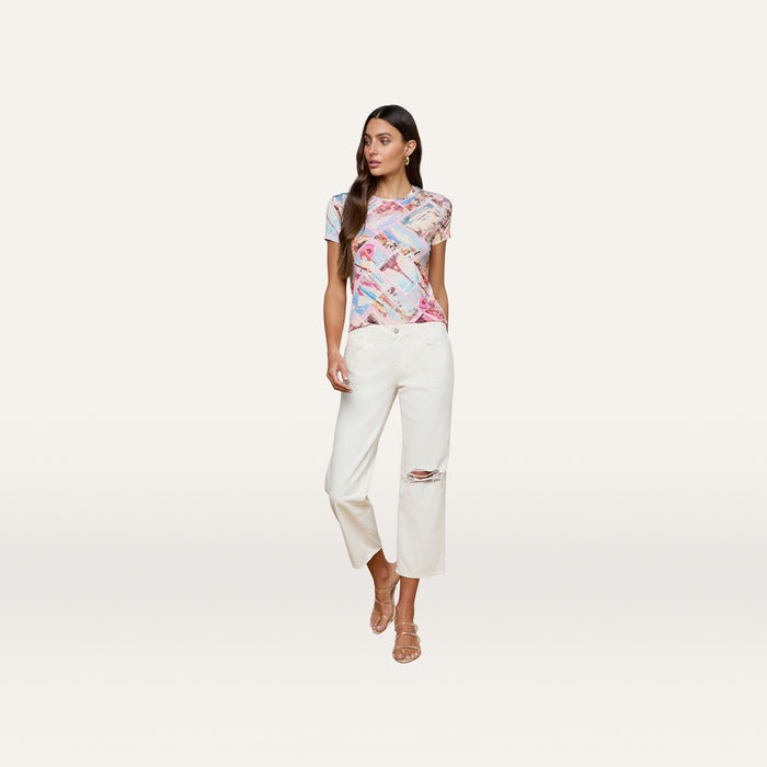 Wearing the bestselling Ressi Fitted Tee in a vibrant print, white wide-leg pants featuring a ripped knee, and beige strappy sandals, the woman stands against a plain white background.