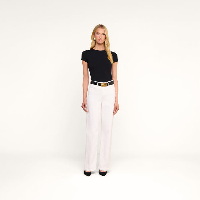 A woman stands facing forward against a plain white background, wearing the Ressi Fitted Tee in black, paired with white high-waisted pants, a black belt with a gold buckle, and black pointed-toe shoes.