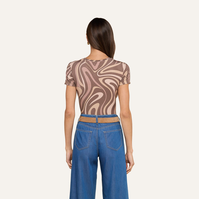 A woman faces away, wearing the bestselling Ressi Fitted Tee featuring an abstract swirl print in brown and beige, paired with high-waisted wide-leg blue jeans. Her straight brown hair falls over her shoulder against a simple off-white background.