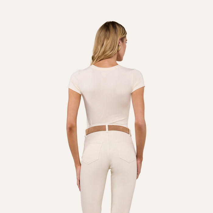 A woman with long blonde hair, wearing the Ressi Fitted Tee in vintage white and high-waisted cream pants with a tan belt, is standing with her back to the camera against a plain background.
