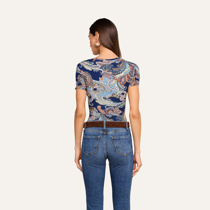 A woman with long brown hair, wearing hoop earrings, models the Ressi Fitted Tee featuring a midnight blue paisley pattern, paired with a brown belt and blue jeans. She is shown from the back against a plain light background.
