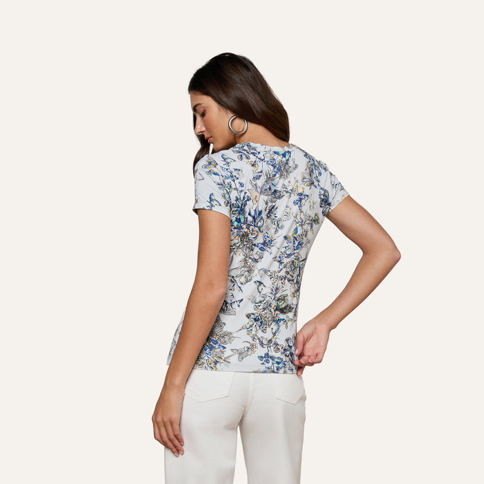 A woman with long brown hair, wearing silver hoop earrings, white pants, and the bestselling Ressi Fitted Tee in a white floral-patterned stretch jersey knit, stands with her back turned and one hand near her lower back against a plain light background.