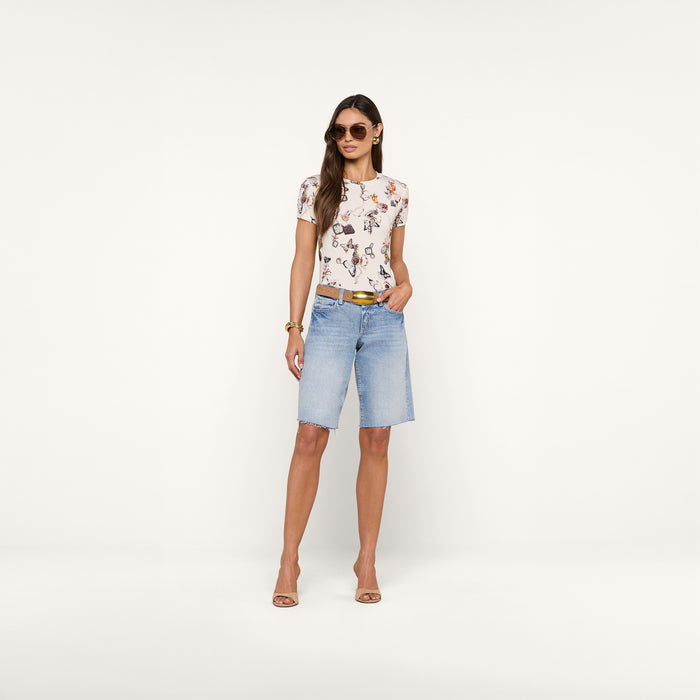 A woman wearing sunglasses showcases the Ressi Fitted Tee in white floral stretch jersey knit with a slim cut, paired with light blue knee-length denim shorts, beige heels, and gold accessories, standing against a plain white background.