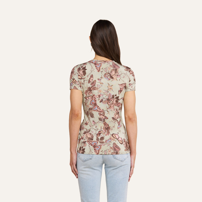 A woman with long brown hair, wearing the beige floral-patterned Ressi Fitted Tee and light blue jeans, stands with her back to the camera against a plain light background.
