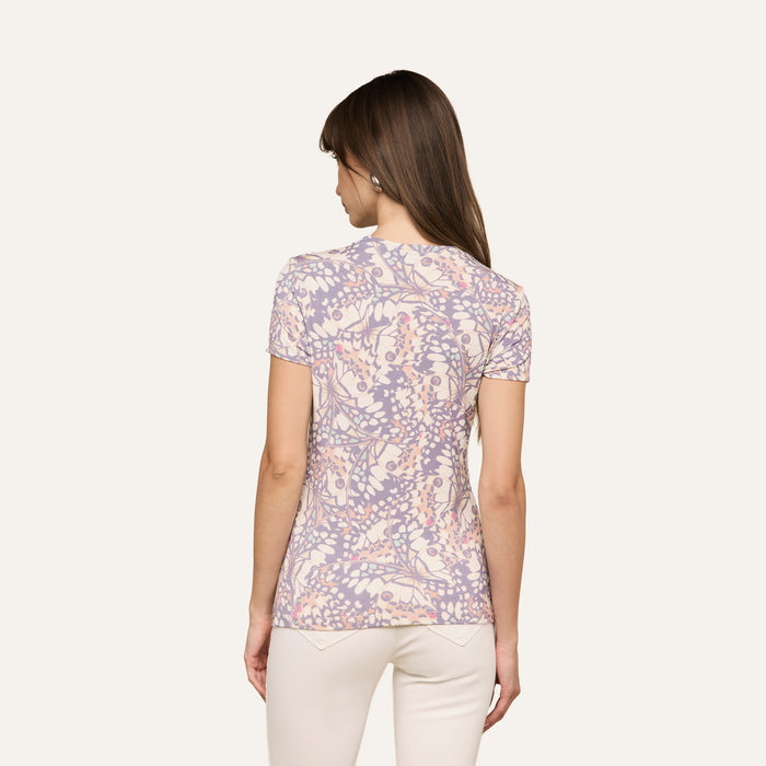 A woman with straight brown hair is standing with her back to the camera, wearing the Ressi Fitted Tee—a short-sleeved, stretch jersey knit top featuring a floral pattern in pastel colors—paired with light-colored pants against a plain background.
