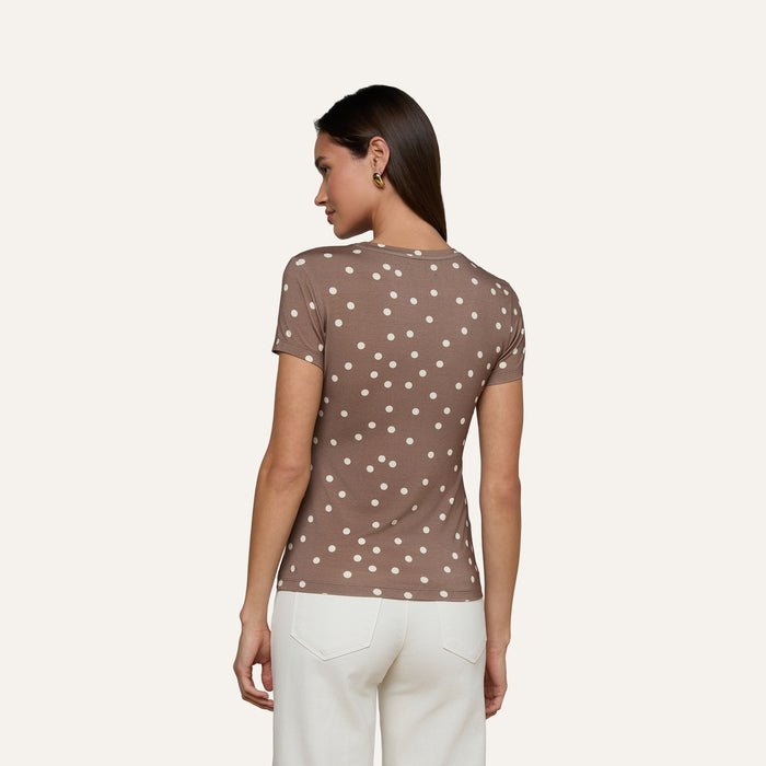 A woman stands facing away, wearing the Ressi Fitted Tee—a slim cut brown short-sleeve shirt with white polka dots—and white pants, set against a plain light background.
