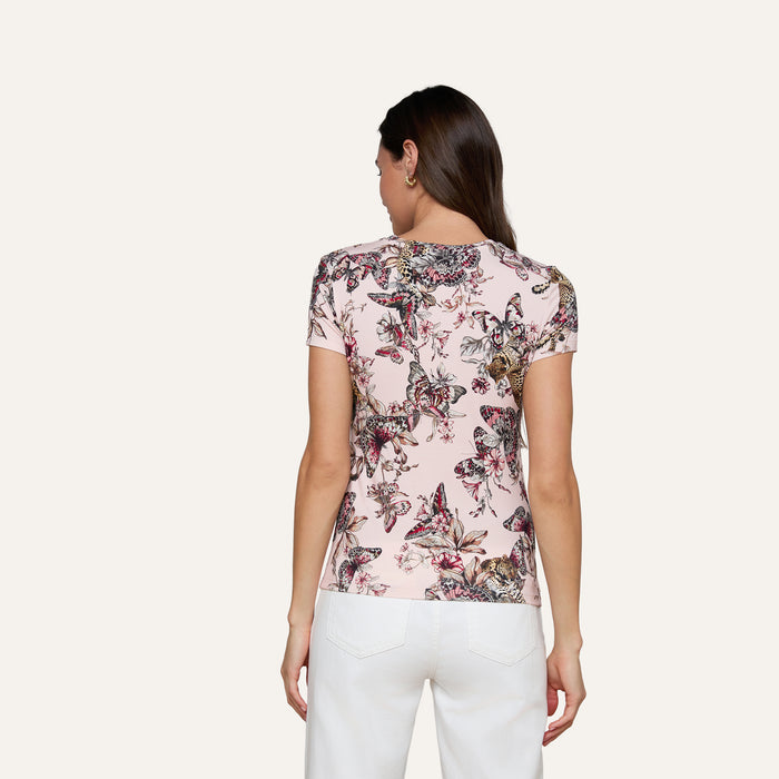 A woman with long brown hair, wearing the Ressi Fitted Tee—a light pink stretch jersey knit short-sleeve shirt with a butterfly jungle print—and white pants, stands facing away against a plain light background.