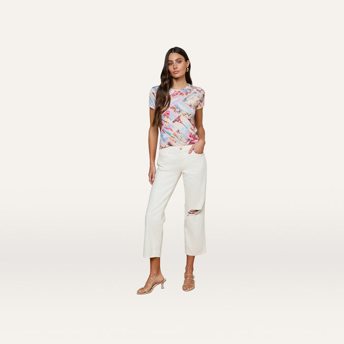 A woman stands against a white background, wearing the bestselling Ressi Fitted Tee in stretch jersey knit, paired with white cropped pants featuring a rip at one knee and beige heeled sandals.