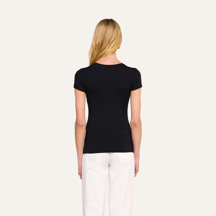A woman with long blonde hair, wearing the Ressi Fitted Tee in black and white pants, stands facing away from the camera against a plain light background.