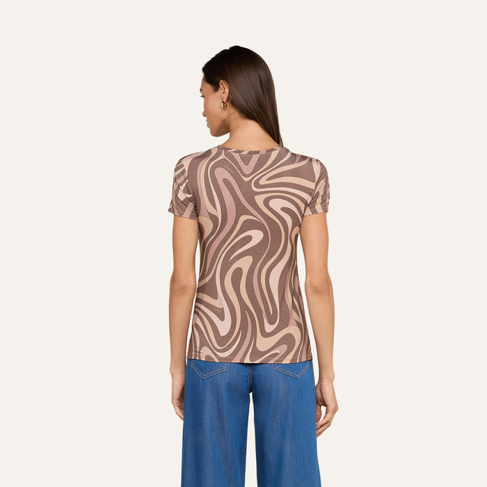 A woman stands facing away, wearing the bestselling Ressi Fitted Tee featuring an abstract swirl print in brown and beige. She pairs it with wide-leg blue jeans. Her straight hair is down, and she has small hoop earrings. The background is plain and light-colored.